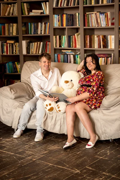 Mujer embarazada y sonriente marido leyendo libro a osito de peluche en —  Fotos de Stock