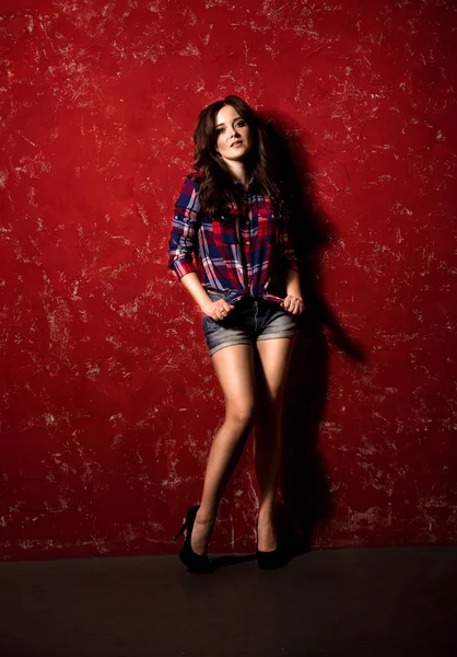 Sexy woman in shorts and checkered shirt posing against red wall — Stock Photo, Image