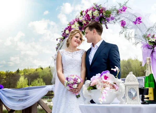 Sposa e sposo in piedi sotto arco floreale nella giornata di sole — Foto Stock