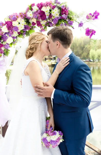 Jonge bruid en bruidegom kussen onder boog op huwelijksceremonie — Stockfoto