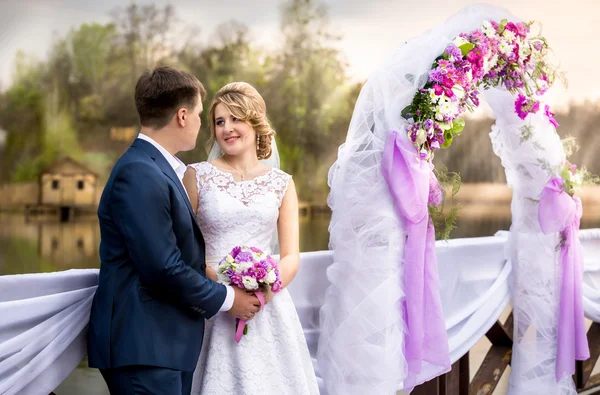 Hermosos recién casados sonrientes posando en el arco floral al atardecer — Foto de Stock