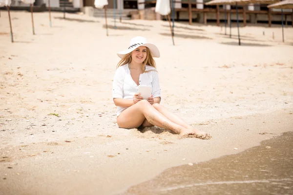 Młoda kobieta w kapeluszu siedzi na plaży i przy użyciu cyfrowego tabletu — Zdjęcie stockowe