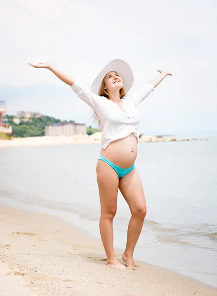 Smiling pregnant woman standing on beach and holding hands up — Stock Photo, Image