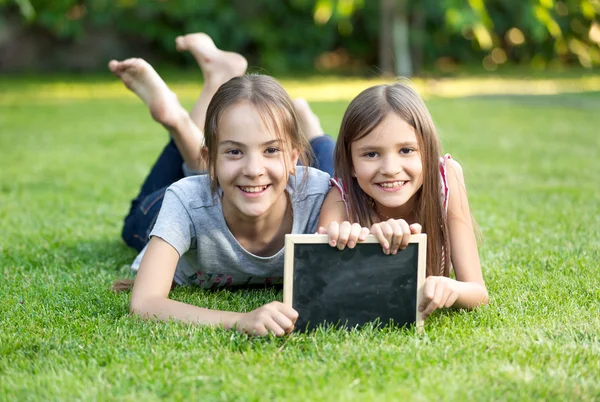 Retrato de duas meninas deitadas na grama com quadro-negro — Fotografia de Stock