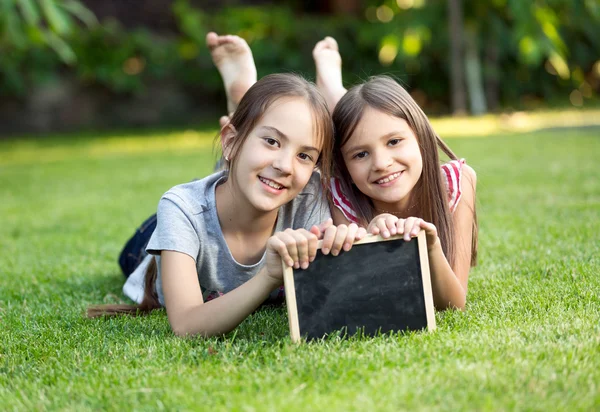 Portret van twee lachende meisjes liggen op gras met schoolbord — Stockfoto