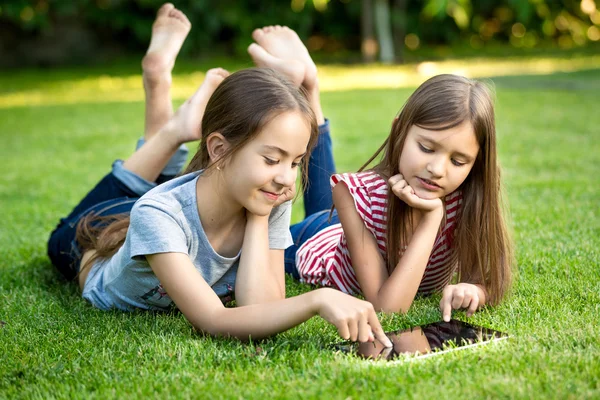 Two sisters lying on grass outdoors and playing on digital table — Stock Photo, Image
