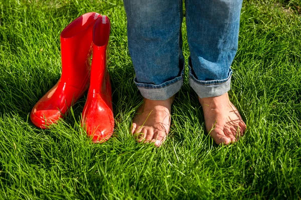 Descalço menina pé próximo vermelho jardim gumboots na grama — Fotografia de Stock