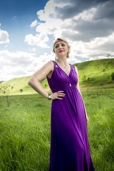 Žena v dlouhé fialové šaty pózuje na louce na slunečný den — Stock fotografie