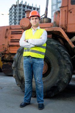 Foreman in helmet posing next to bulldozer with crossed hands