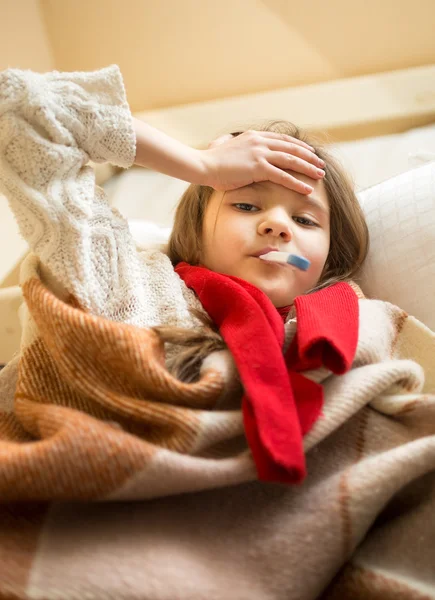 Маленька дівчинка в шарфі лежить в ліжку і вимірює температуру — стокове фото