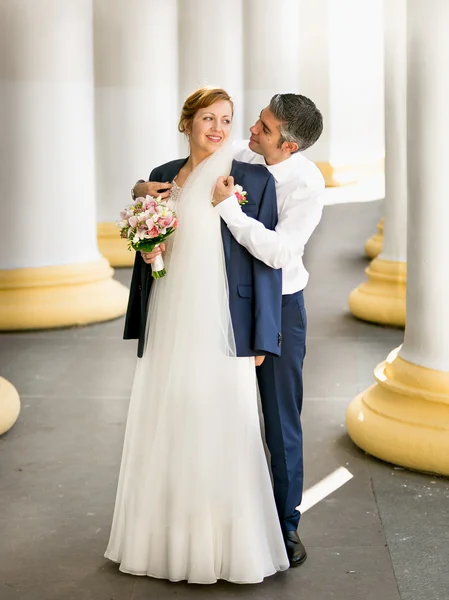 Young groom hugging beautiful bride from back at ancient columns — Stock Photo, Image