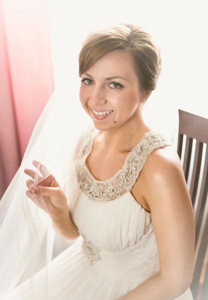 Smiling bride sitting on chair next to window at bedroom — Stok fotoğraf