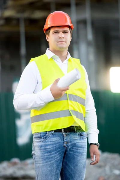 Architect in hardhat posing with blueprints against scaffolding — Stock Photo, Image