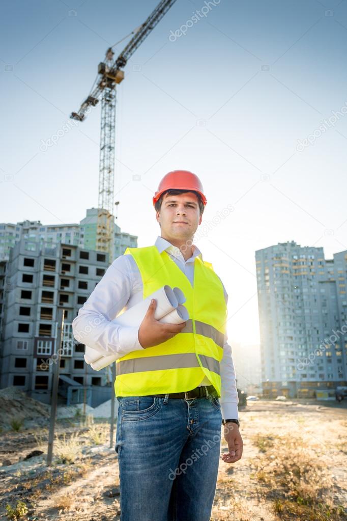 engineer in hardhat and yellow jacket posing with blueprints