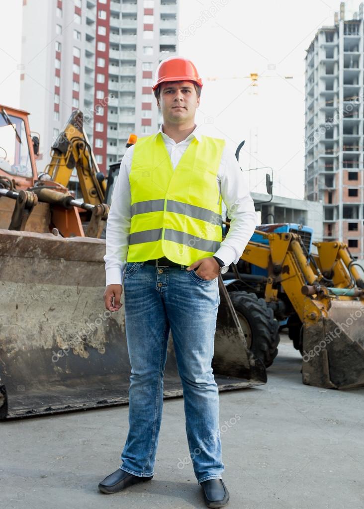Businessman in safety jacket and hardhat posing next to bulldoze