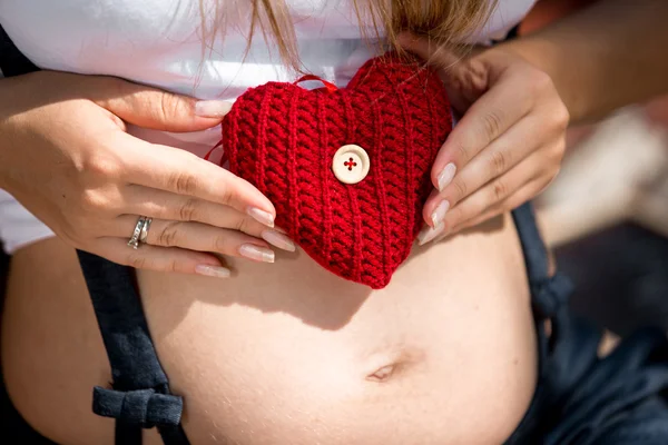 Închiderea unei tinere gravide care ține inima roșie pe abdomen — Fotografie, imagine de stoc