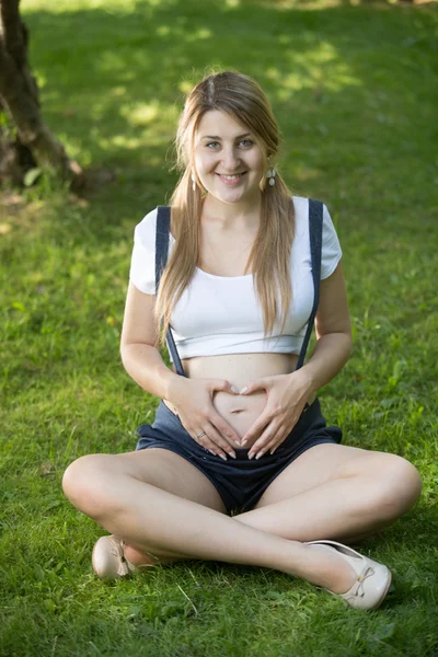 Gravid kvinna avkopplande på park och hålla händerna på buken — Stockfoto