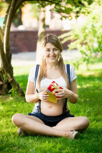 Park çimenlerin üzerinde oturan ve hediye kutusu tutan hamile kadın — Stok fotoğraf