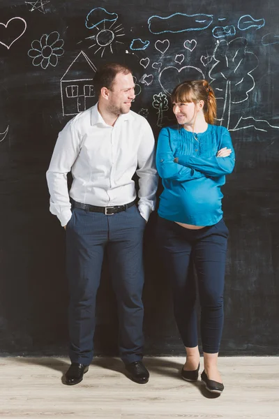 Laughing husband and pregnant wife posing against big blackboard — Stock Photo, Image