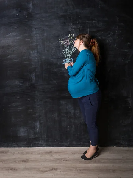 Pregnant woman smelling flowers drawn by chalk on big black wall — Stock Photo, Image