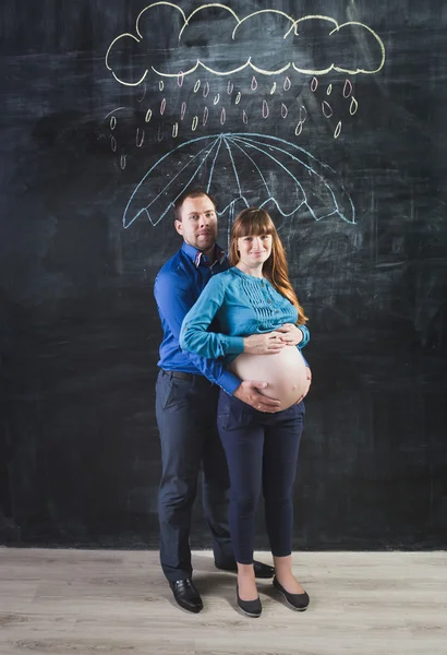 Husband hugging pregnant woman under umbrella at rain against ch — Stock Photo, Image