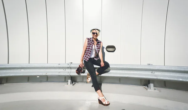 Smiling hipster girl sitting on metal fence at parking lot — Stock Photo, Image