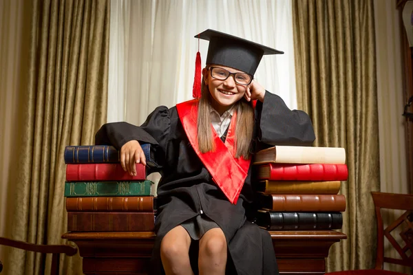 Menina em boné de formatura sentado na mesa e inclinando-se sobre livros — Fotografia de Stock