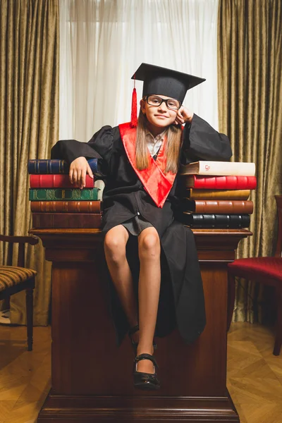 Kleines Mädchen mit Abschlussmütze posiert auf Tisch in der Bibliothek — Stockfoto