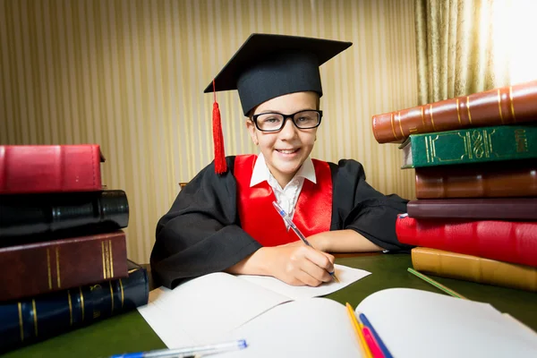 Chica sonriente en gorra de graduación sentada en la mesa entre pila de —  Fotos de Stock