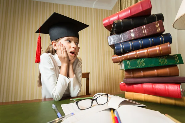 Girl in graduation cap looking at high heap of books on table at — Stock Photo, Image