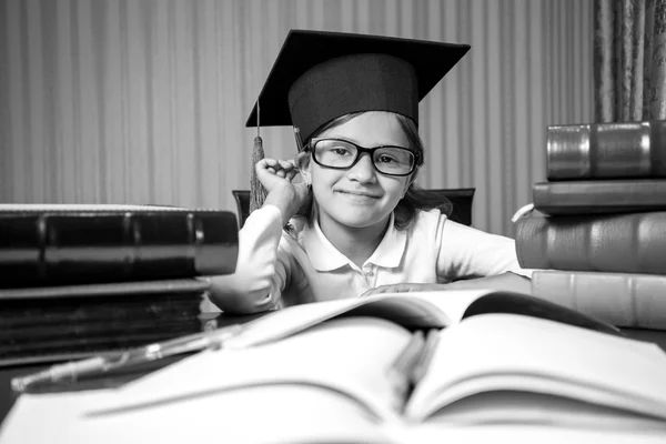 Portrait of smart girl in graduation hat sitting at desk full of — Stock Photo, Image