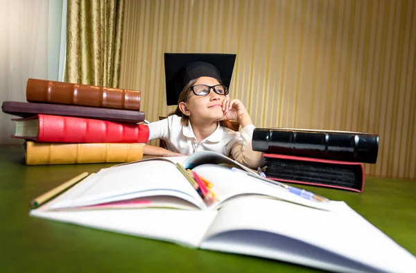 Thoughtful girl in graduation cap dreaming while sitting at tabl — Stock Photo, Image