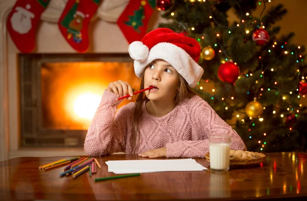 Nachdenkliches Mädchen in Weihnachtsmütze denkt an Brief mit Geschenkwünschen — Stockfoto