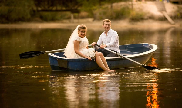 Happy bride and groom sitting in wooden rowing boat at sunset — Stock Photo, Image