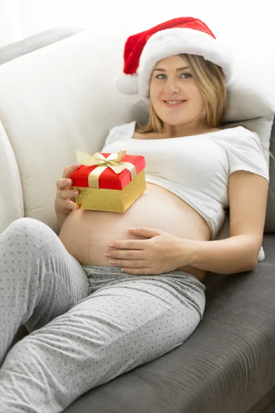 En lykkelig, gravid kvinne i nisselue som ligger på sofaen med gaveboks – stockfoto