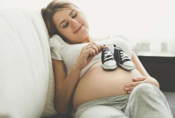 Glücklich schwangere Frau liegt auf Sofa und hält Babystiefel — Stockfoto