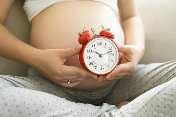 Fecho da mulher grávida segurando despertador vermelho na barriga — Fotografia de Stock