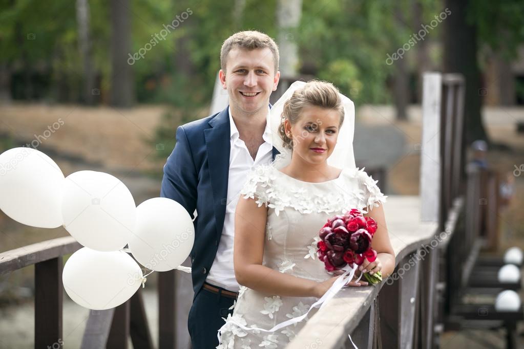happy smiling newlyweds posing with white balloons on the bridge