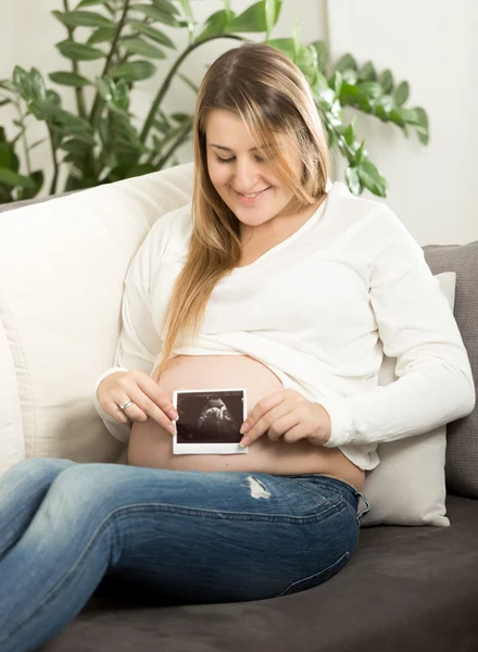 Smiling pregnant woman sitting on sofa and holding ultrasound fe — Stock Photo, Image