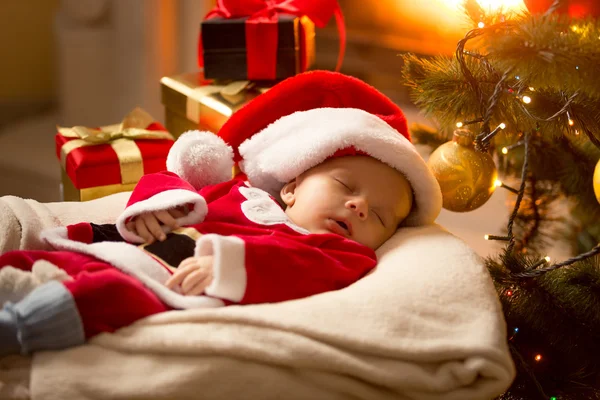 クリスマス ツリーの横にある暖炉のそばで眠っているサンタ衣装で男の子の赤ちゃん — ストック写真