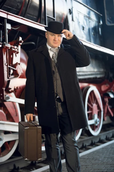 Человек в ретро-костюме, шляпа-котелок и чемодан, позирующий на старом локомоте — стоковое фото
