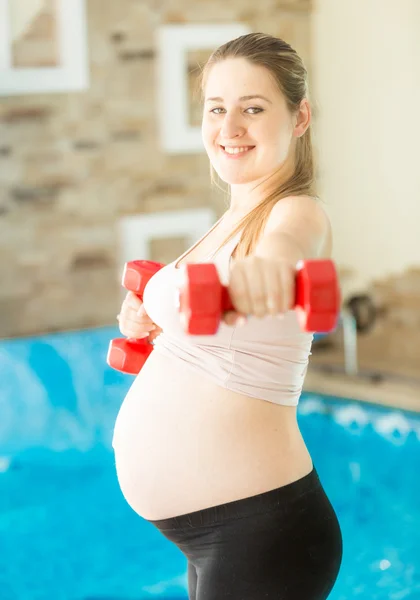 Mujer embarazada sonriente haciendo ejercicio con pesas en el gimnasio — Foto de Stock