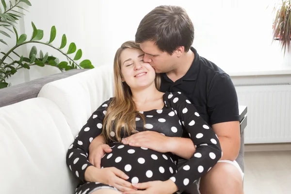 Pohledný muž sedí na pohovce a líbání těhotná žena v tvář — Stock fotografie