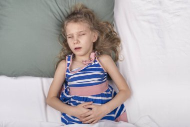 Stüdyo ışıkları. Okul çağındaki bir kız yatakta yatar ve karnına tutunur. Yakın plan.