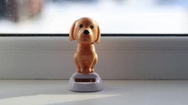 Doğal ışık. Kahverengi bir oyuncak köpek beyaz bir pencerenin üzerinde duruyor ve başını sallıyor. Güneşten güç alıyor.