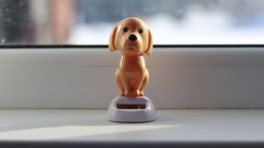 Doğal ışık. Kahverengi bir oyuncak köpek beyaz bir pencerenin üzerinde duruyor ve başını sallıyor. Güneşten güç alıyor.