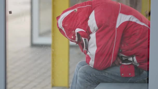 Un hombre irreconocible con una chaqueta roja se sienta en una parada de transporte público. Sin cara. — Vídeos de Stock