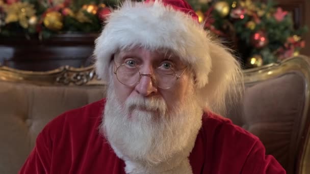 Добрий і веселий Санта Клаус сидить на стільці, дивлячись на фотокамеру, і налаштовує свої окуляри і посміхається. Портрет добрих нових років Санта. Портрет Святого Миколая з білою бородою на Різдво — стокове відео