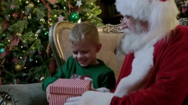 Uzun gri sakallı yaşlı adam Noel Baba hediye kutusundaki çocuğa, kutudan parlak ışık saçan küçük sarışın çocuğa şömineli sandalyede oturan ve Noel ağacını odada süsleyen.