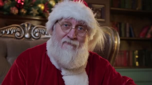 Père Noël mignon regarde opérateur de caméra et sourit joyeusement. Portrait genre souriant Père Noël avec barbe grise en costume et lunettes d'or assis dans la chaise sur fond de décorations de Noël. — Video